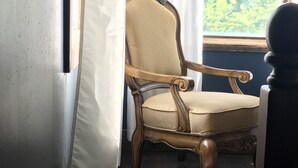 Ein Zimmer mit einem Holzstuhl neben einem Fenster mit Vorhängen, einem dunklen Holzboden und Blick ins Grüne.
