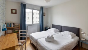 Une chambre d’hôtel avec deux lits, un bureau avec une chaise, une bibliothèque et une fenêtre avec des rideaux.