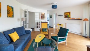 Un salon avec un canapé bleu, une table jaune et un fauteuil vert. Une porte blanche donne accès à une autre pièce.