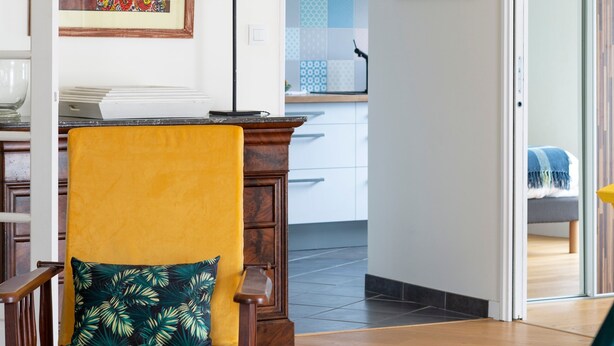 Un salon moderne avec un fauteuil jaune, un canapé vert et un coin repas comprenant une table et des chaises. Au fond, on aperçoit une cuisine avec des meubles blancs et un luminaire bleu au plafond.