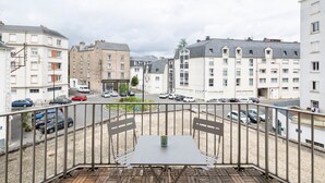 Un balcon avec une table et des chaises, donnant sur une rue bordée de voitures garées et d’immeubles.