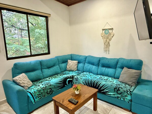 A teal-colored sectional sofa with patterned cushions, a wooden coffee table with a remote control, and a wall-mounted dreamcatcher. - 3 min walking distance to Nosara beach (Guanacaste)