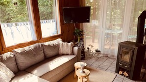 A cozy living room with a beige sectional sofa, a flat-screen TV mounted on the wall, a small wooden stool, and a wood-burning stove.