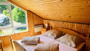 A wooden-paneled bedroom with a bed, pillows, a nightstand, a lamp, and a window overlooking greenery.
