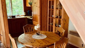 A wooden dining table with six chairs, a small white orchid centerpiece, and a kitchen area with wooden cabinets and a window.