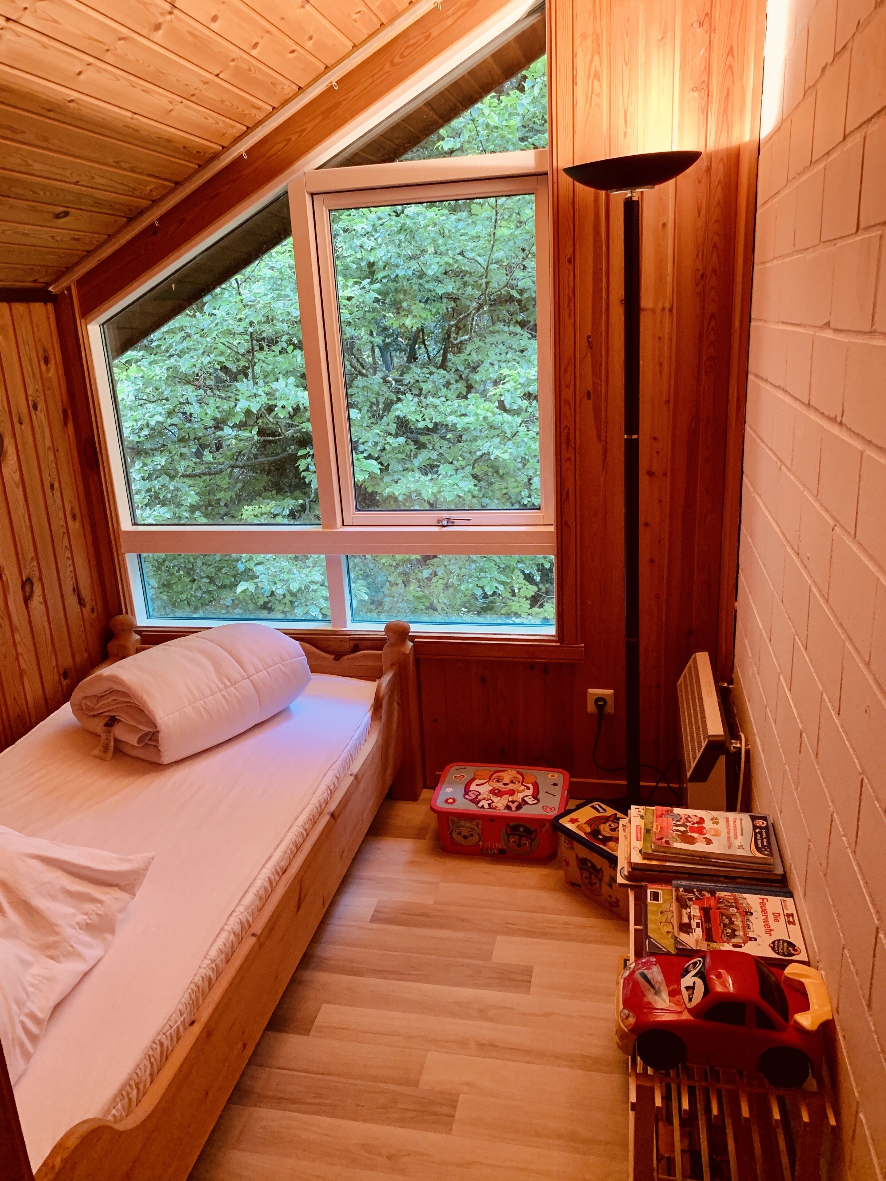 Ein gemütlicher Holzraum mit einem Bett, einem Fenster mit Blick auf Grünflächen sowie einem kleinen Tisch mit Spielzeug und Büchern.