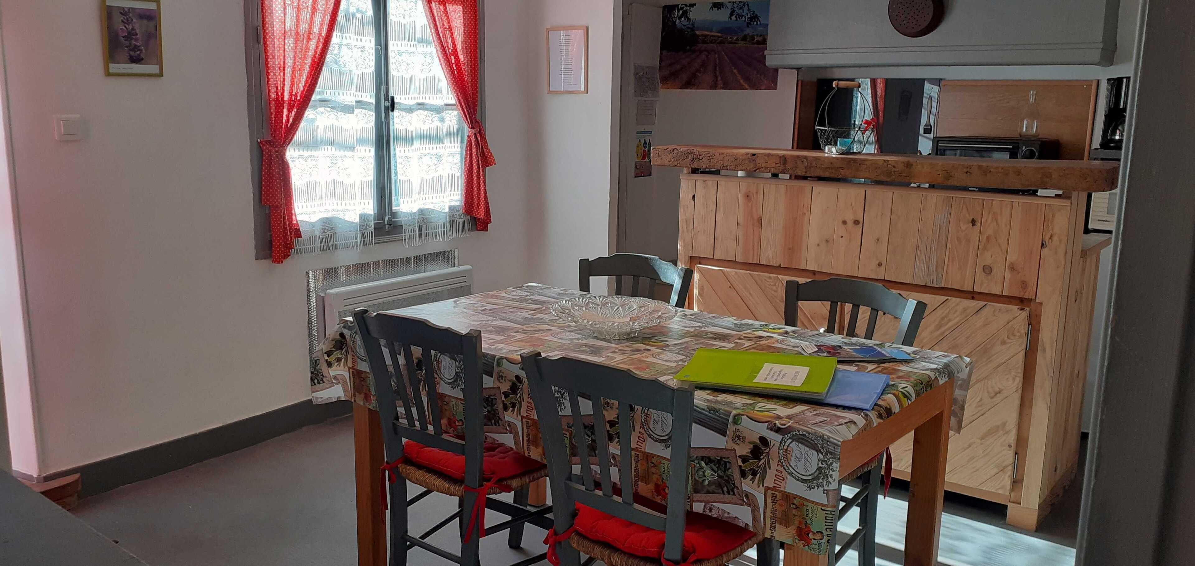 A dining area with a table covered in a patterned tablecloth, surrounded by chairs with red cushions. A wooden cabinet with a countertop is visible in the background.