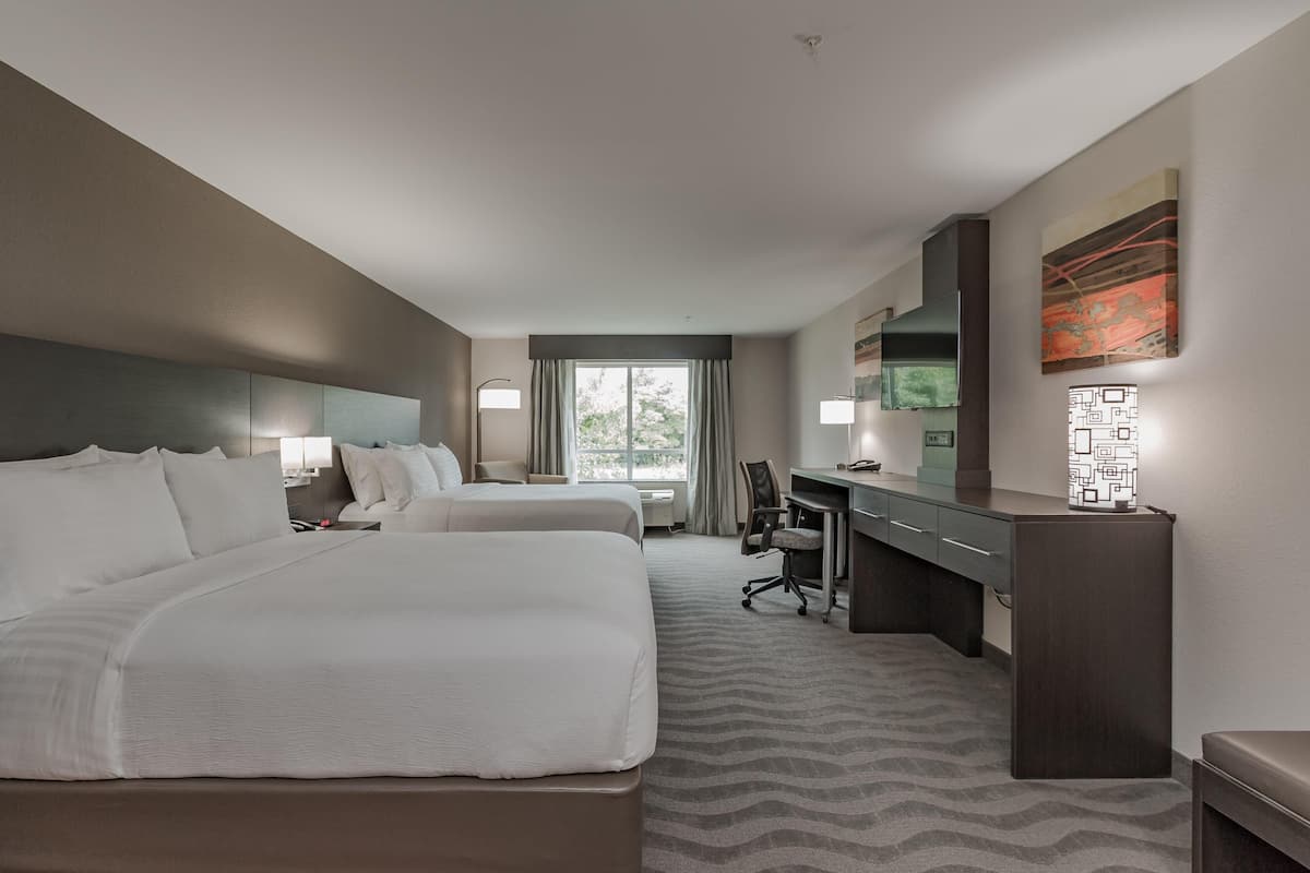 A hotel room with two beds, a desk, a chair, a lamp, a mirror, and a window with curtains.