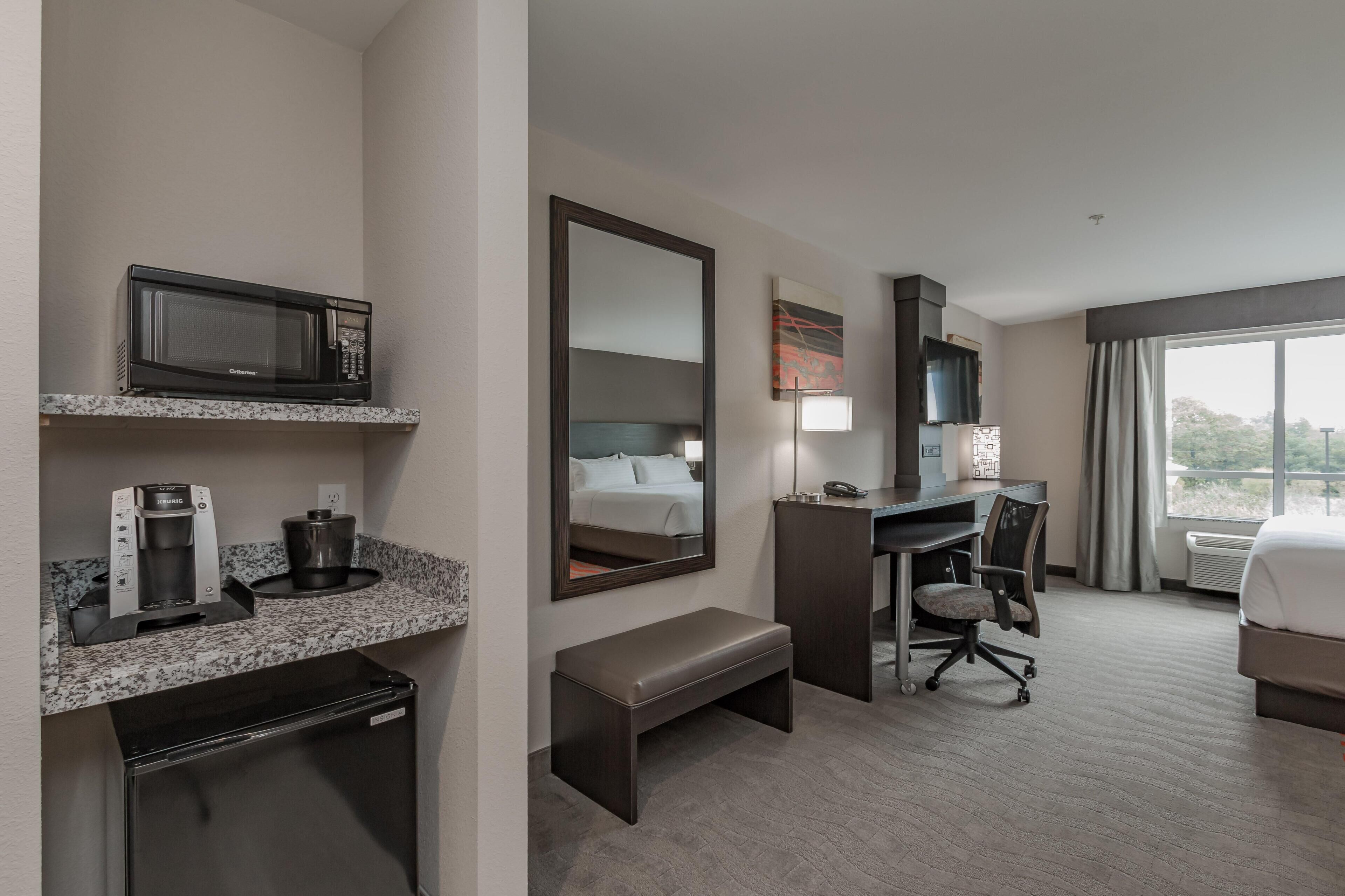 A hotel room with a kitchenette, a bed, a desk, a chair, and a large window.