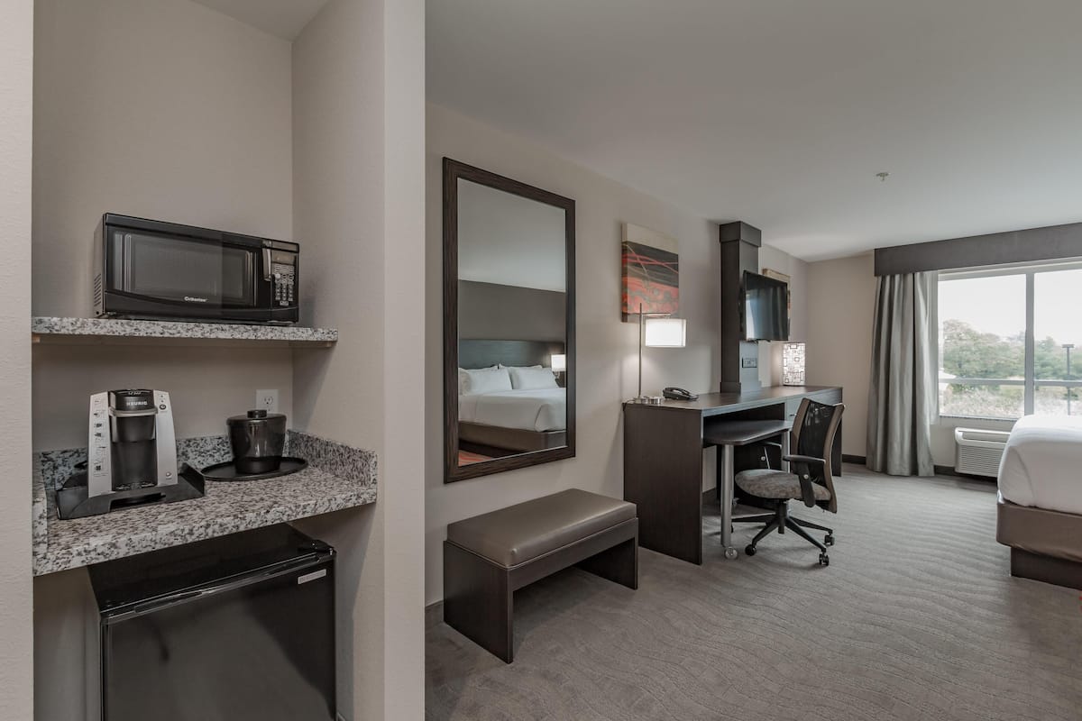 A hotel room with a kitchenette, a bed, a desk, a chair, and a large window.