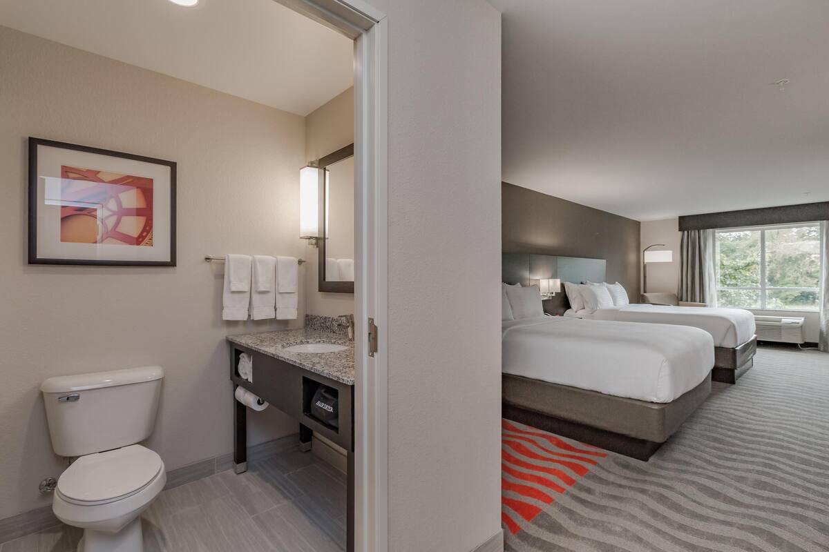 A hotel room with a bathroom, two beds, a large window, and a painting on the wall.