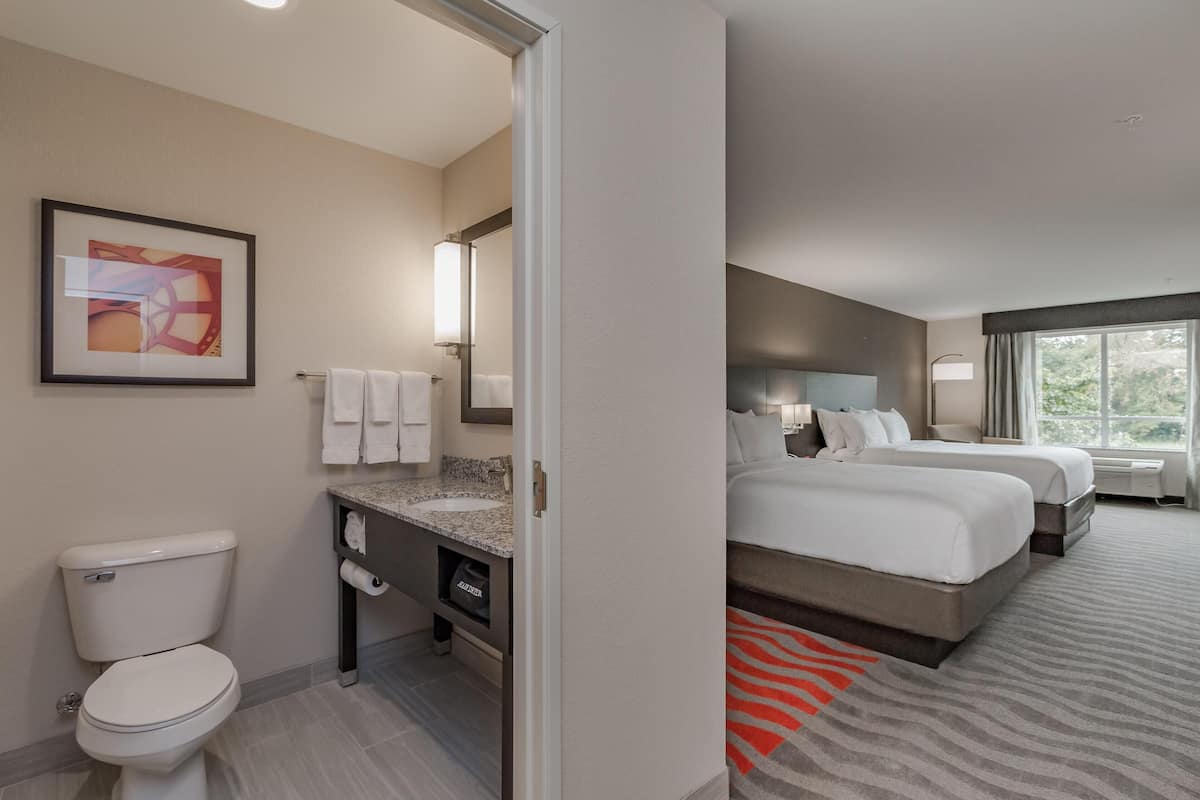 A hotel room with a bathroom, two beds, a large window, and a painting on the wall.