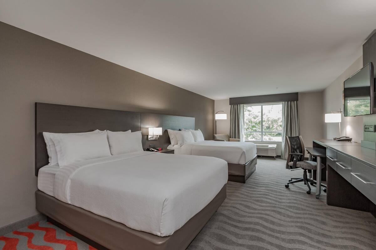 A hotel room with two beds, a desk, a chair, a lamp, and a large window.