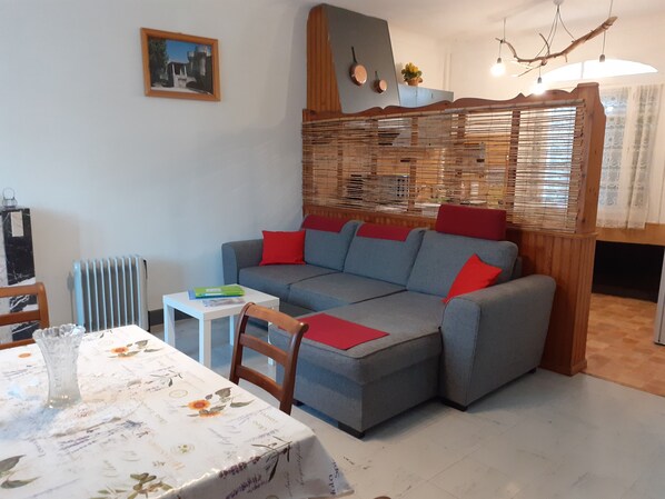 A living room with a grey sofa, red cushions, a white table with a floral tablecloth, and a radiator.
