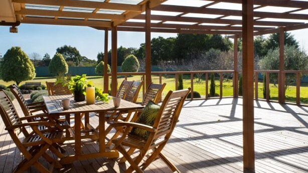 Un espacio cubierto para comer al aire libre, con una mesa y sillas de madera, rodeado por una terraza de madera y vistas a un jardín cuidado.