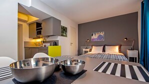 Une chambre d’hôtel moderne dotée d’un coin cuisine, d’une table à manger avec deux bols en métal, d’un lit recouvert d’une courtepointe à carreaux et d’une table de chevet avec une lampe.