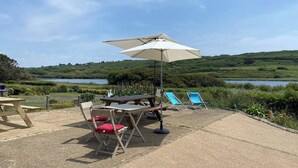 An outdoor seating area with a table and chairs, a large umbrella, and a view of a lake and hills. - Beachfront at Baie des Trépassés (Cléden-Cap-Sizun)