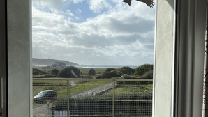 A view from a balcony with a wooden deck, a metal fence, and a distant landscape including hills and a lighthouse. - Beachfront at Baie des Trépassés (Cléden-Cap-Sizun)