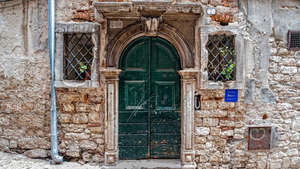 Un edificio storico con una doppia porta verde, pareti in pietra e una piccola finestra con inferriata.
