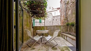 Un piccolo patio esterno con un tavolo rotondo e due sedie, un pavimento in pietra e vista su un edificio bianco con una persiana verde.