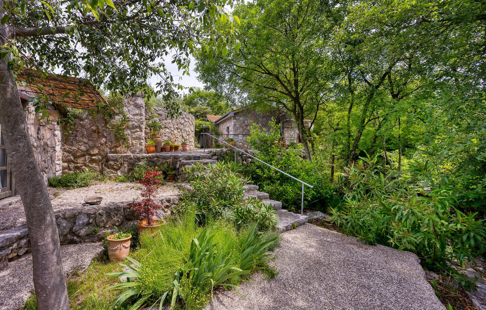 A rustic stone building with a stone staircase leading to a wooden deck, surrounded by lush greenery and potted plants.