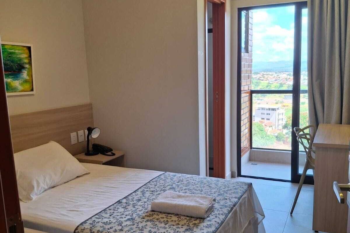 Habitación de hotel con cama, escritorio, silla, equipo de aire acondicionado y vista de la ciudad a través de un ventanal grande.