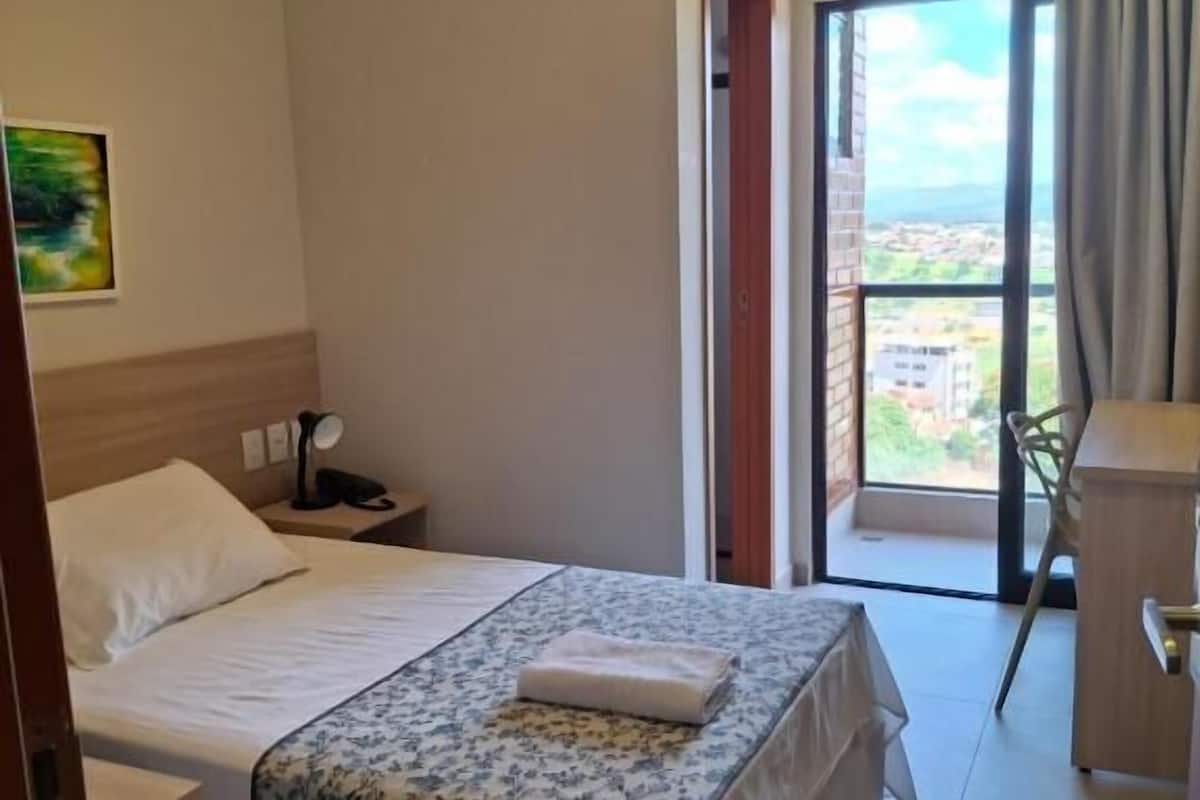 Habitación de hotel con cama, escritorio, silla, aire acondicionado y vistas al exterior.