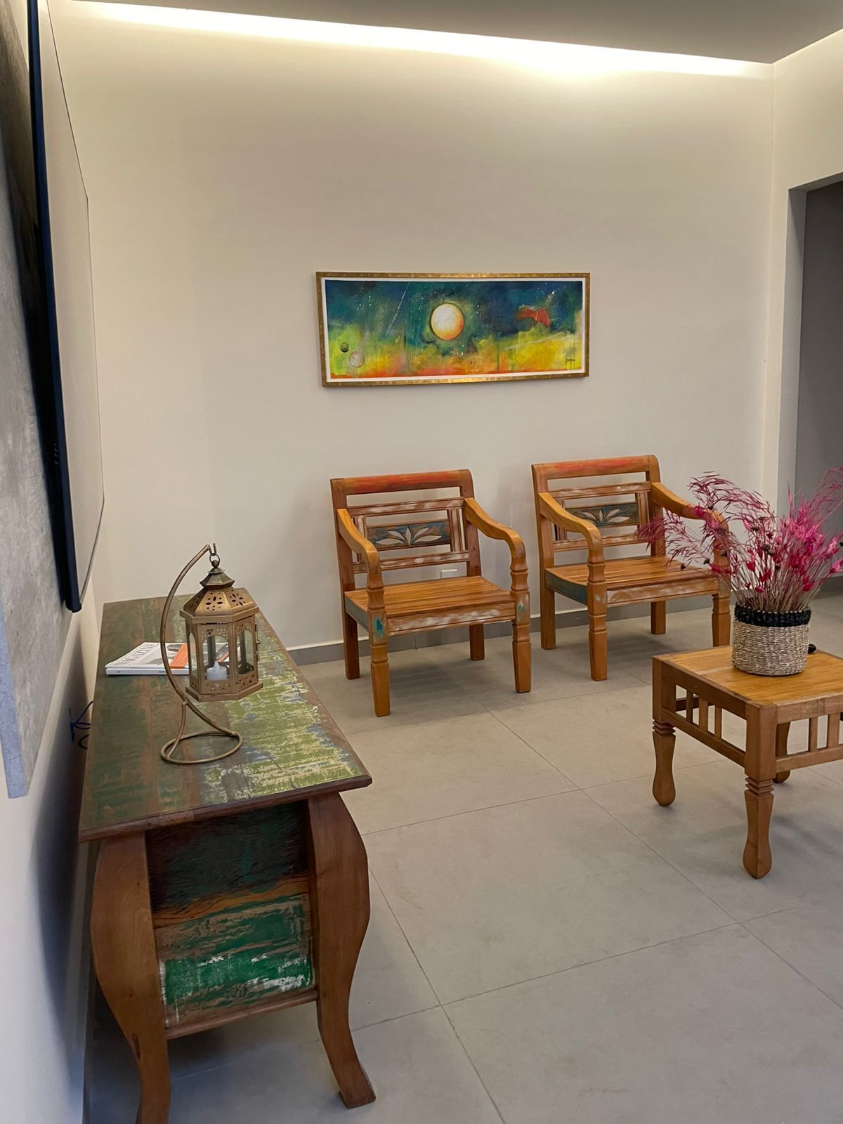 Una habitación con un escritorio de madera, dos sillas, una mesita con un florero y un cuadro en la pared.
