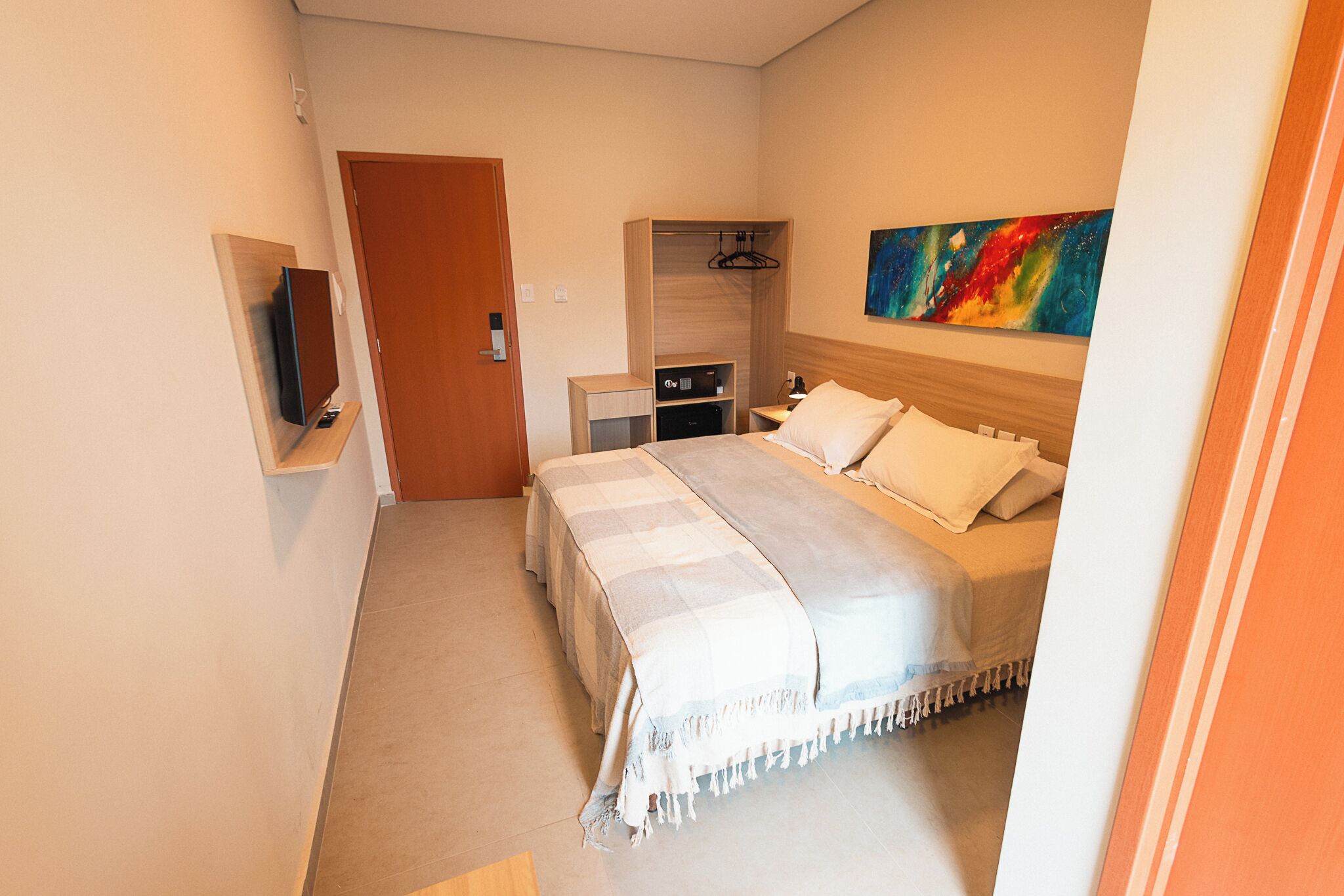 Quarto de hotel com cama, mesas de cabeceira, televisão, guarda-roupa e um quadro na parede.