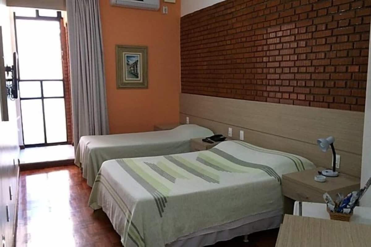 Una habitación con dos camas, una pared de ladrillo, una puerta con cortinas y una pequeña mesa con lámpara.