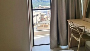 A hotel room with a bed, a desk, and a chair. There is a window with curtains, and a view of buildings and mountains outside. - Hotel Verdes Mares (Ouro Branco)