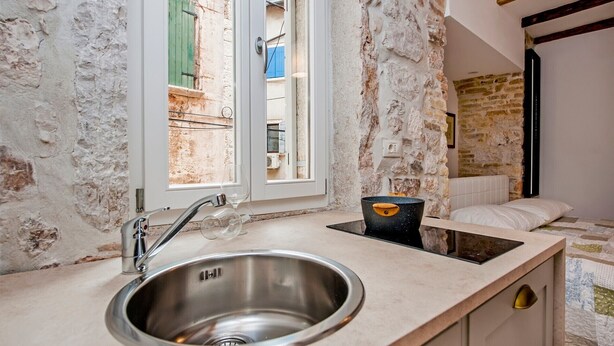 Una cocina con un lavamanos de acero inoxidable, una ventana con contraventanas verdes y una pared de piedra.