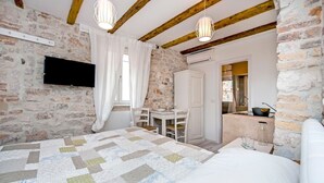 Un dormitorio con pared de piedra, vigas de madera, una cama, un televisor, un escritorio y un baño visible a través de una puerta abierta.