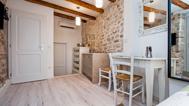 Een knus kamertje met een stenen muur, een witte deur, een houten plafond, een wit bureau met stoel, en een spiegel.