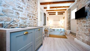 Een kleine kamer met een stenen muur, een houten plafond, een bed, een bureau en een aan de muur bevestigde televisie.