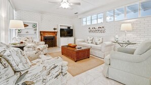 A spacious living room with white brick walls, a fireplace, a flat-screen TV, and a large window. - Beautiful East Beach Cottage with Pool (St. Simons)