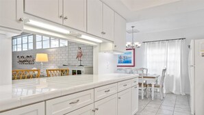 A modern kitchen with white cabinetry, a large island, and a dining area with a white table and chairs. - Beautiful East Beach Cottage with Pool (St. Simons)