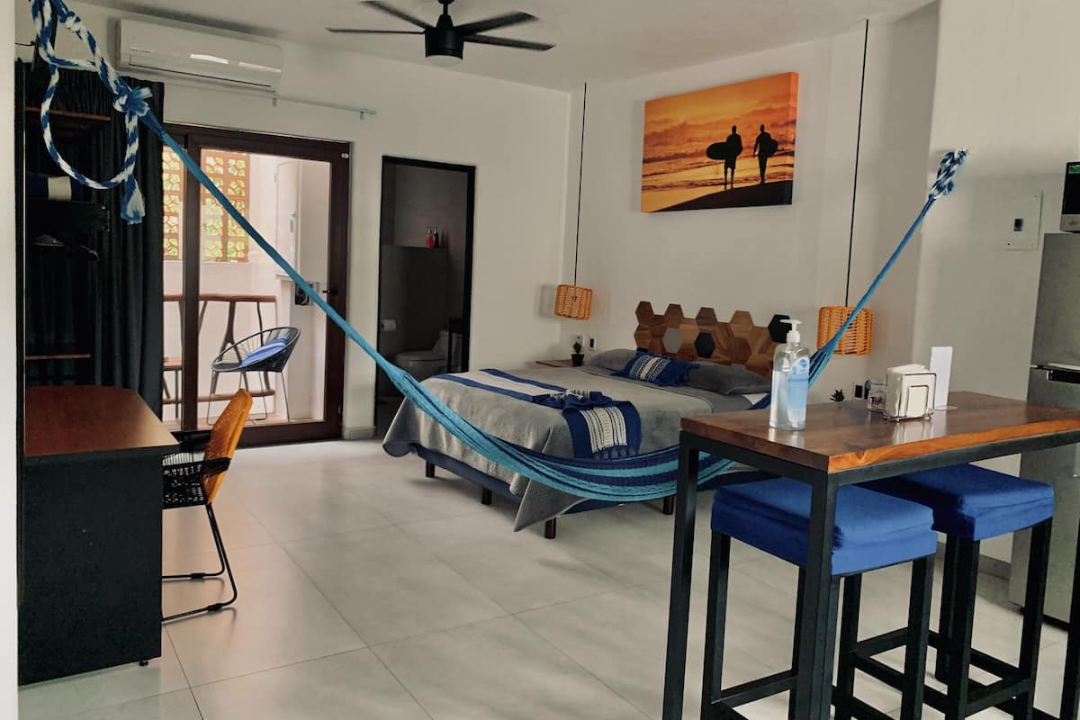 Un dormitorio con cama, escritorio, silla, hamaca y un cuadro en la pared.