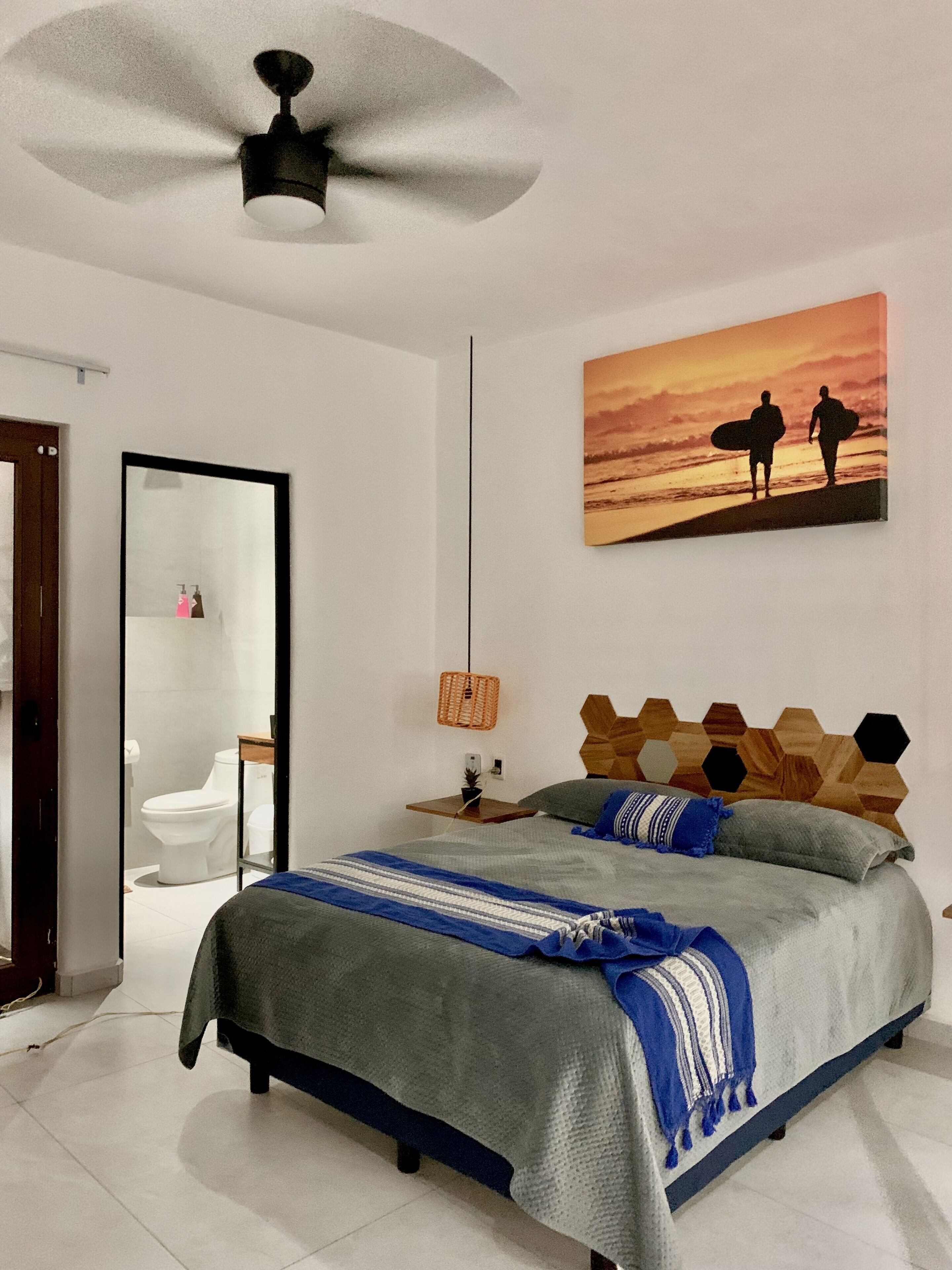 Un dormitorio con cama, ventilador de techo, cuadro de surfistas, cabecero de madera y mesita de noche con lámpara.