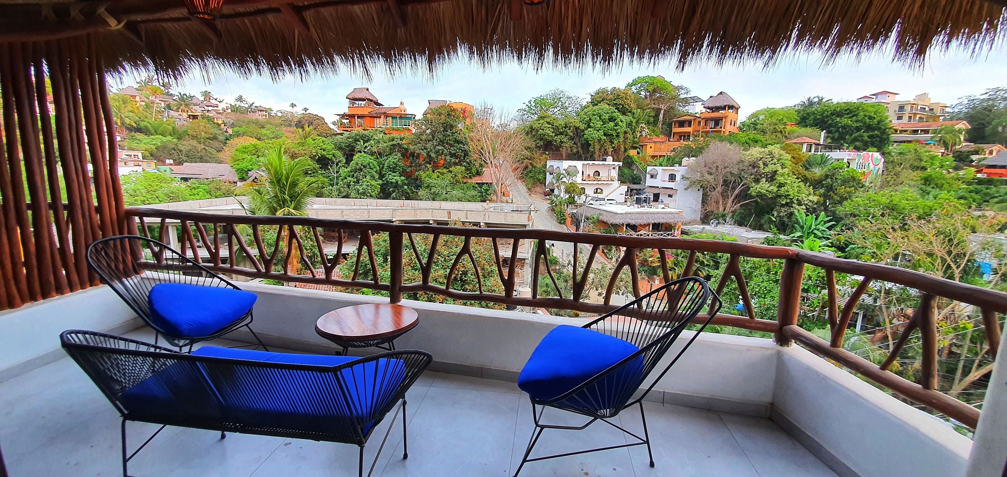 Un balcone con due poltrone blu imbottite e un piccolo tavolino rotondo, con vista su una collina con diversi edifici e vegetazione lussureggiante.
