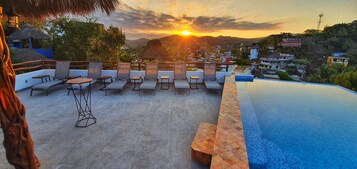 Un área de piscina en la azotea con sillones de descanso y vistas a la puesta de sol sobre un paisaje urbano.