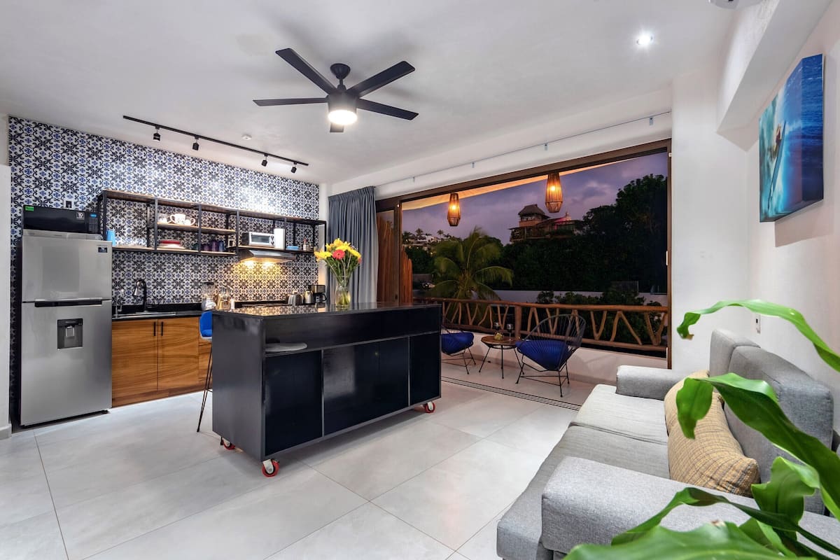 Una cocina moderna con una isla negra, un comedor con sillas azules y un ventanal con vistas a palmeras y al atardecer.