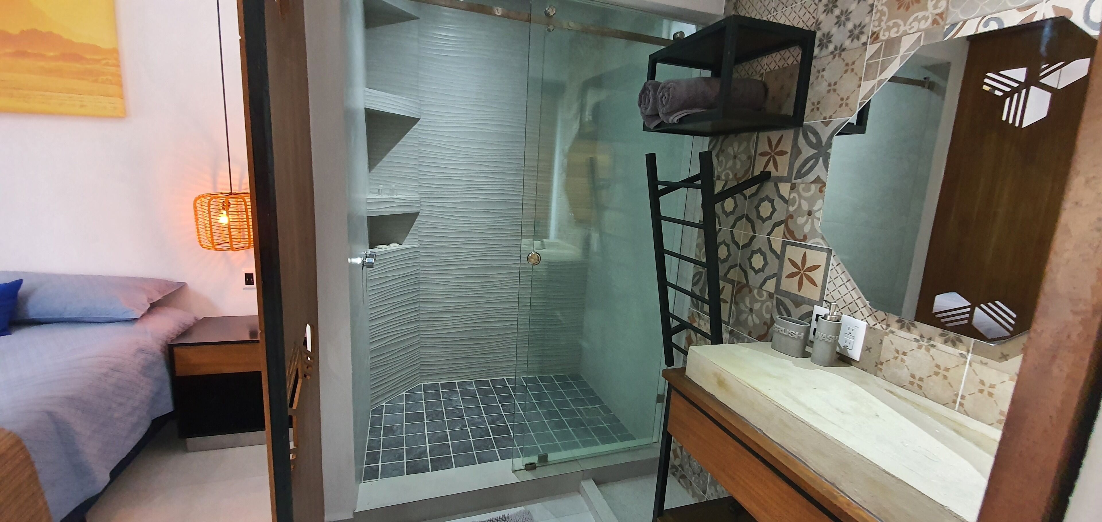 A bathroom with a glass shower enclosure, a wooden vanity, and a bed with a blue pillow.