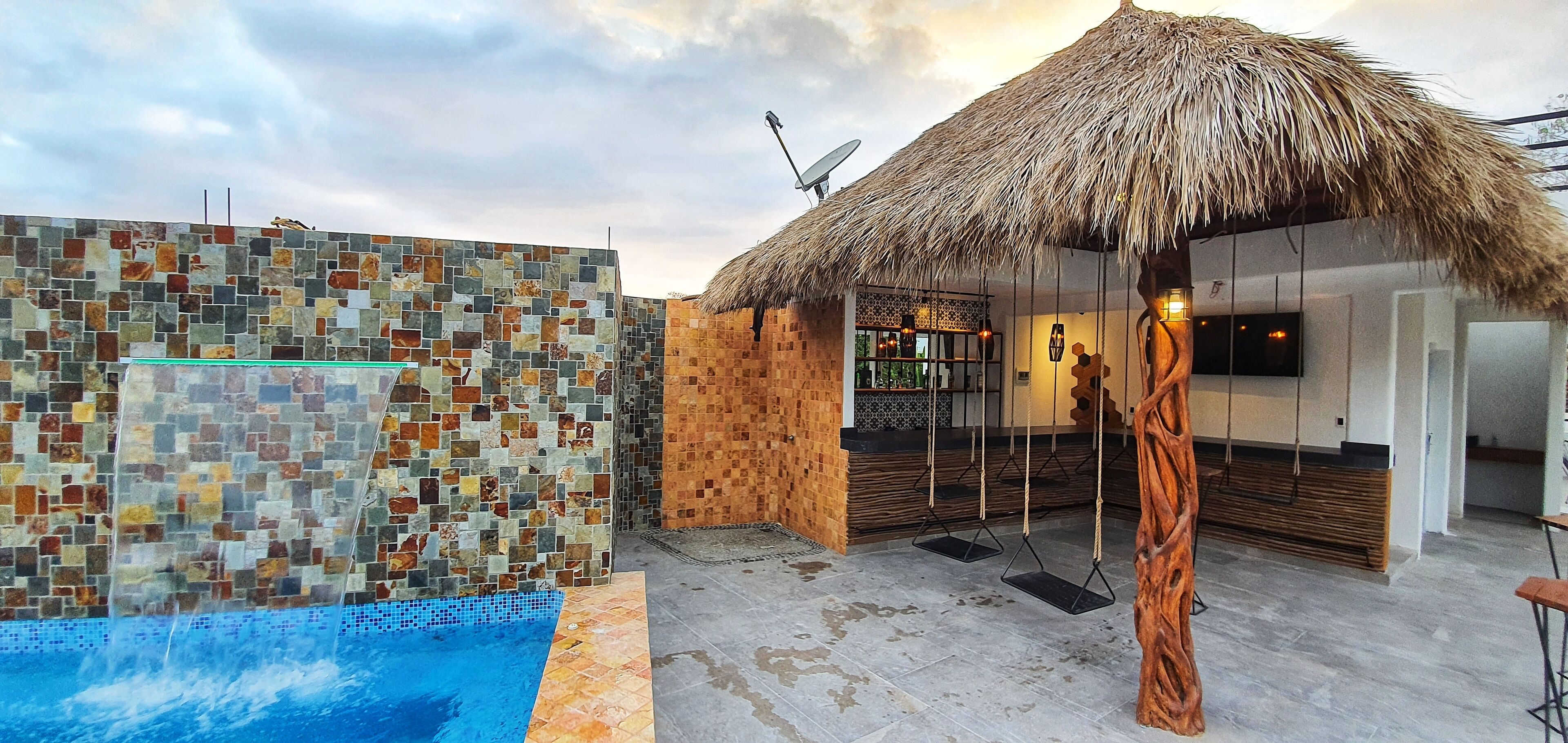 A pool area with a mosaic wall, a thatched-roof structure, and a wooden bench.