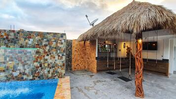 A pool area with a mosaic wall, a thatched-roof structure, and a wooden bench.