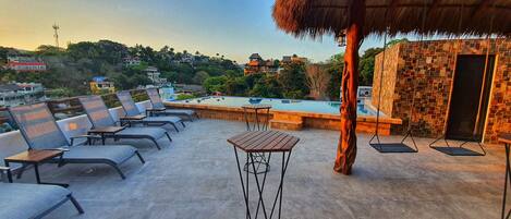 A rooftop terrace with a swimming pool, lounge chairs, and a thatched-roof structure.