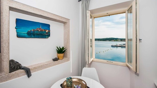 A room with a view of a harbor, a framed painting of a coastal scene, a potted plant, and a small table with decorative items. - Studio Sunrise with Sea view (Rovinj)