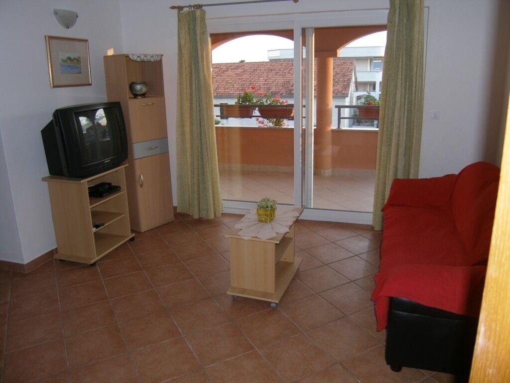 Una sala de estar con un televisor sobre un mueble de madera, un sofá rojo y un ventanal corredizo que da a un balcón con plantas en macetas.