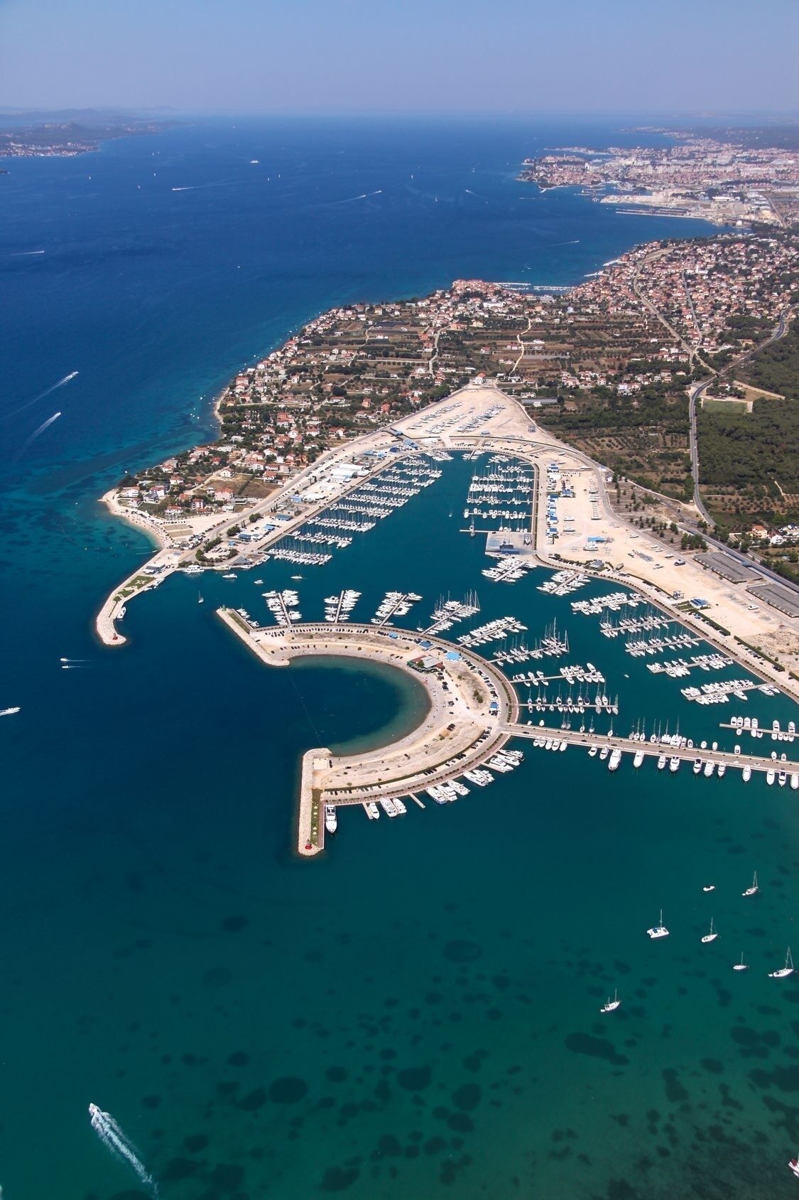 An aerial view of a marina with numerous boats docked, surrounded by a coastal town with visible roads and buildings.