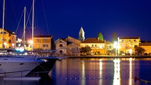 Un port de plaisance de nuit, avec des bateaux amarrés, une église dotée d’un clocher et des bâtiments dont les lumières se reflètent sur l’eau.